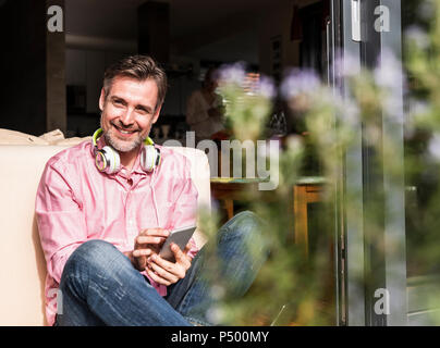 Portrait of smiling mature man sitting at open terrace door using  smartphone Stock Photo