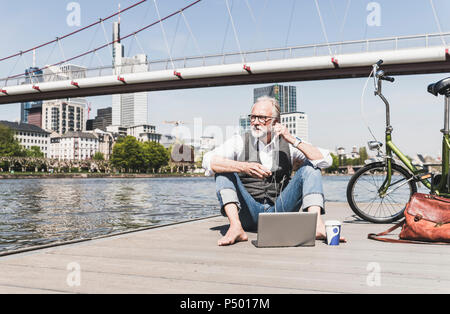 Mature man with laptop, earbuds and bicycle sitting at the riverside in the city Stock Photo