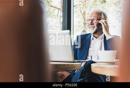Mature businessman sitting at table in a cafe using cell phone and laptop Stock Photo