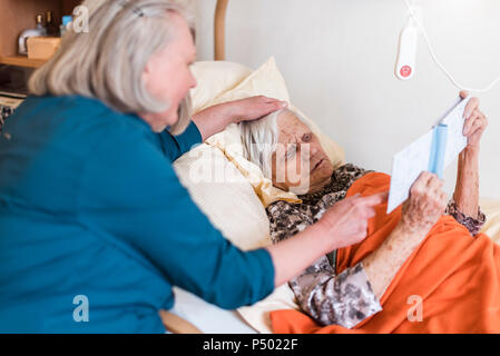 Woman taking care of old woman lying in bed reading book Stock Photo
