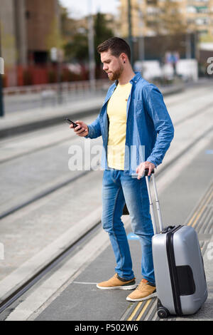 Young man waiting at a station with smartphone in his hand and trolley Stock Photo