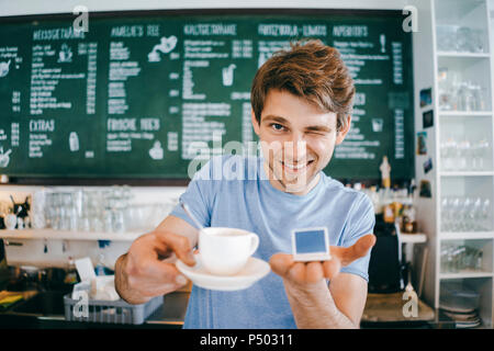 Smiling man in a cafe offering cup of coffee and holding miniature laptop model Stock Photo