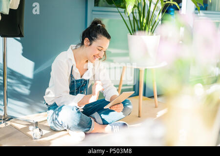 Smiling young freelancer sitting on the floor in her studio using tablet Stock Photo