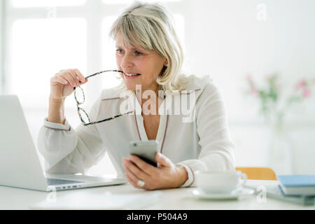 Mature businesswoman holding cell phone working on laptop at desk Stock Photo