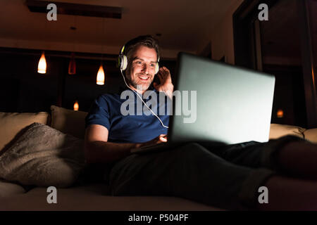 Man with headphones sitting on couch at home looking at laptop Stock Photo