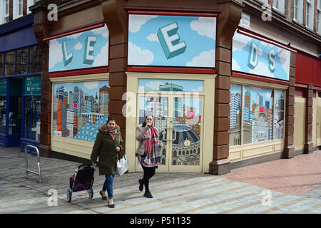 Two young women walk past an empty storefront with a wallpaper illustration of the city of Leeds, in a pedestrian zone, Leeds England, United Kingdom. Stock Photo