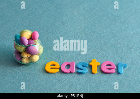 A small glass container filled with colorful speckled eggs and the word easter on a blue background Stock Photo