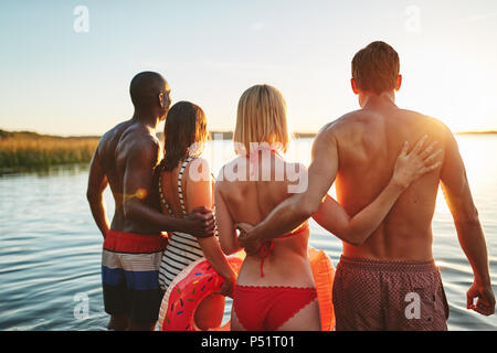 Rearview of two young couples in swimsuits watching the sunset while standing arm in arm together in a lake Stock Photo