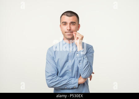 Young latin man in blue shirt keeps fore finger on lips, tries to keep conspiracy. Try to keep secrets. Isolated shot of attractive man on gray backgr Stock Photo