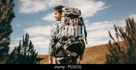Rear view shot of happy young man with backpack going on a camping. Caucasian male hiker on mountain looking away and smiling. Stock Photo