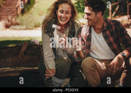 Smiling young camping couple eating roasted sweets. Man and woman having roasted marshmallows on camping. Stock Photo