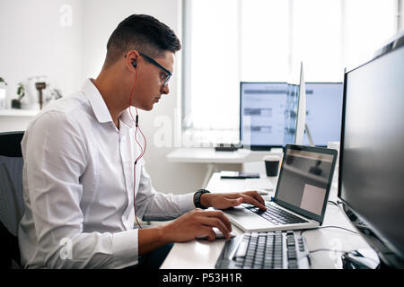 Software developer sitting at his office desk working on laptop wearing earphones. Man wearing spectacles working on laptop computer in office. Stock Photo