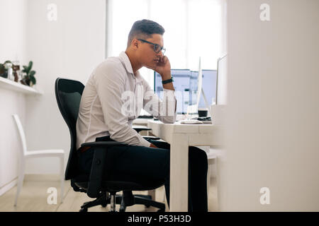 Software developer sitting at his office desk looking at the computer and thinking with hand on cheek. Man wearing spectacles working on computer in o Stock Photo