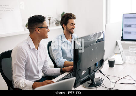 Software developers sitting at office working on computers. Smiling application developers talking while working on computers in office. Stock Photo