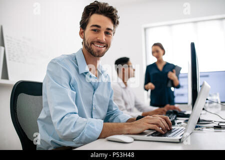 Software developers sitting at office working on computers. Happy application developer working on a laptop in office with his colleagues in the backg Stock Photo