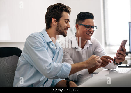 Application developers testing application on mobile phone in  office. Smiling office colleagues sitting at office desk looking at a mobile phone. Stock Photo