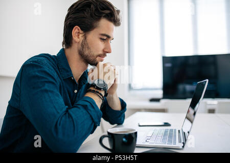 Software developer sitting in front of computer and working in office. Man looking at laptop sitting with his chin resting on hands with a coffee cup  Stock Photo