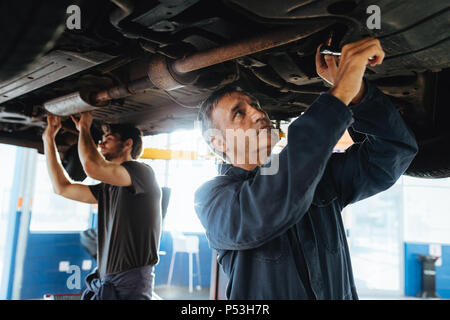 Two mechanics working under a car. Men in garage repairing exhaust system of a lifted automobile. Stock Photo