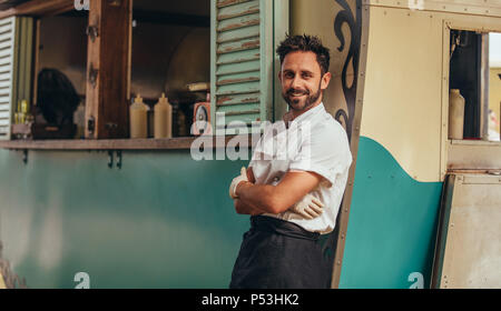 Portrait of happy young salesman standing by his food truck. Proud food truck owner standing with his arms crossed and looking at camera. Stock Photo
