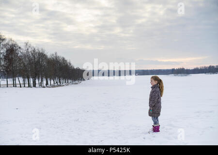 Little blonde caucasian Swedish girl standing outdoor in winter landscape. Beautiful cold evening at sunset with snow on field and tree alley Stock Photo