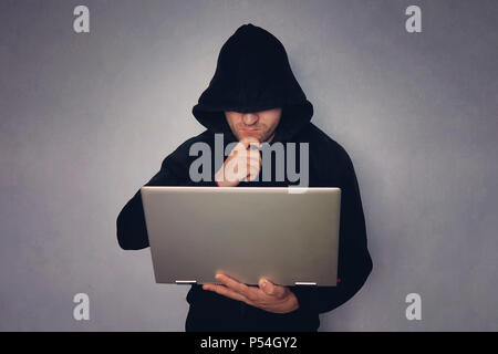 cybercrime, thoughtful hacking and technology crime - male hacker in dark room with laptop computer, hacker in a black hood with a gray modern ultrabo Stock Photo