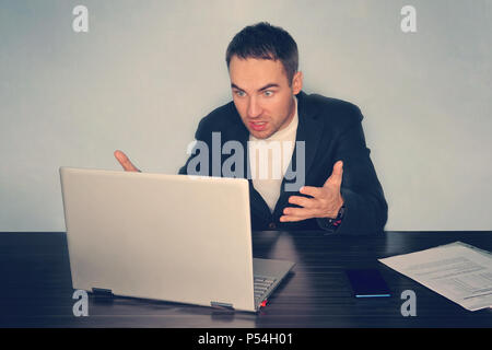 Frustrated young businessman looks at laptop screen screaming in anger at workplace in office. Unexpected high bill, unpaid debt, failing financial re Stock Photo