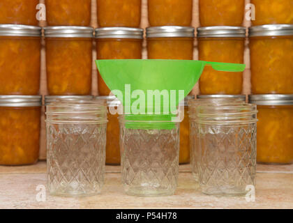 Green funnel sitting in empty canning jars waiting to be filled on kitchen counter. Finished jars of peach jam stacked in rows in background. Stock Photo