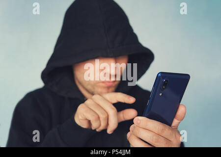 Faceless unrecognizable hooded person using mobile phone, identity theft and technology crime concept, selective focus on body. hacking a smartphone.  Stock Photo