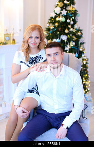 Young wife sitting with husband wearing white shirt near Christmas tree. Stock Photo