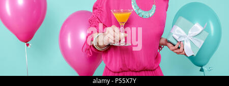 Gorgeous young woman in party outfit holding birthday present and cocktail, isolated over pastel blue colored background. Birthday Party banner. Stock Photo