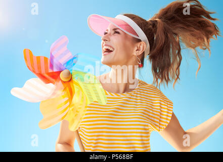 cheerful healthy woman in yellow shirt against blue sky playing with colorful windmill Stock Photo