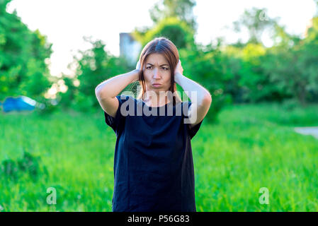 A girl in summer in park in black T-shirt. Beautiful asian. He covers his ears with his hands. Emotionally loud noise. Headache. Unwillingness to listen to interlocutor. Something terrible happened. Stock Photo
