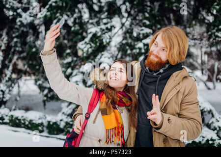 Young couple in love rest in park winter. A man hugs girlfriend, woman holds mobile phone in her hand and takes photo of herself. People use smartphon Stock Photo