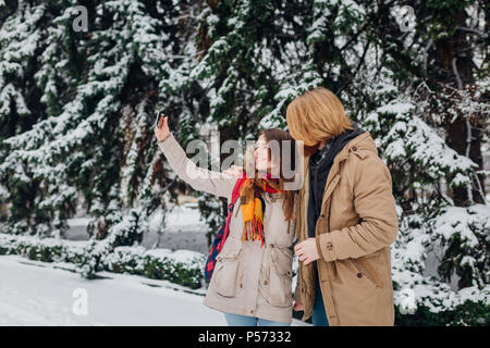 Young couple in love rest in park winter. A man hugs girlfriend, woman holds mobile phone in her hand and takes photo of herself. People use smartphon Stock Photo