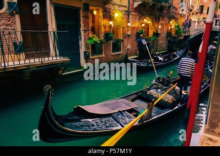 evening assembly of gondolas with gondolieres in a narrow canal in the antique heart of venice with shining lanterns and emerald green water rear view Stock Photo