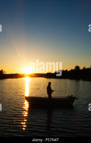 Silhouette image of a man standing and fishing in a rowing boat in a small lake at sunset. Stock Photo