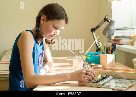 Teenage girl at home is engaged in creativity, draws watercolor at a table in room. Stock Photo