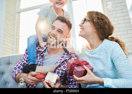 Warm toned portrait of playful happy family holding presents siting on sofa in living room at home, focus on smiling handsome man Stock Photo