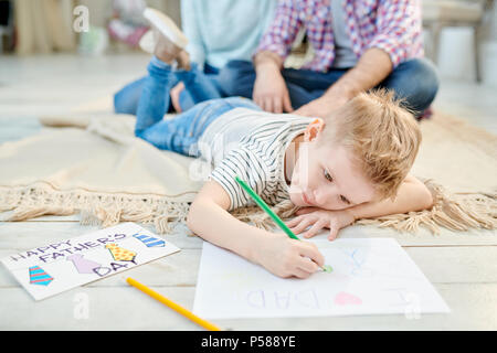 Warm toned portrait of cute little boy lying on floor in living room drawing picture for Fathers day for dad and smiling, copy space Stock Photo