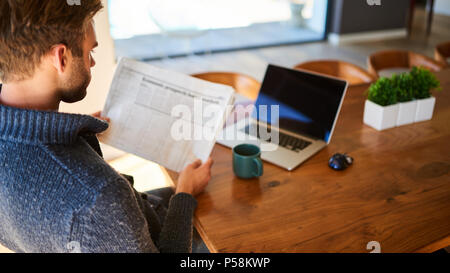 over the shoulder image of an attractive young man holding a newspaper while seated at his beautiful wooden dining table at home, with a hot cup of tea and his laptop ready to work. Stock Photo