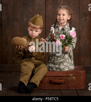 children boy are dressed as soldier in retro military uniforms and girl in pink dress sitting on old suitcase, dark wood background, retro style Stock Photo