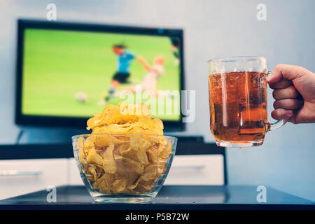 Beer mug with beer in hand a man next to the chips on the background of a large TV on the screen which is a football match at the world Cup. soccer me Stock Photo