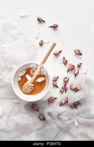 Flat lay of honey in small ceramic bowl, wooden honey dipper, silk, petals and dried roses on a white background with copy space. Top view. Stock Photo