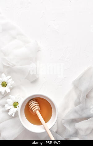 Flat lay of honey in small ceramic bowl, wooden honey dipper, flowers, petals on a white background with copy space. Top view. Stock Photo