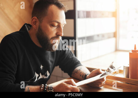 Young tattooed man holding menu at cafe, side view. Stock Photo