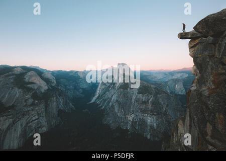 A fearless hiker is standing on an overhanging rock enjoying the view towards famous Half Dome at Glacier Point overlook in beautiful post sunset twil Stock Photo