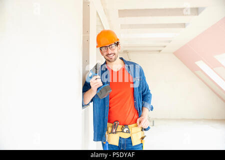 Portrait of young repairman wearing safety helmet and tool belt and using hammer drill driver while working on construction site. Stock Photo