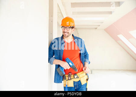 Portrait of young repairman wearing safety helmet and tool belt and using hammer drill driver while working on construction site. Stock Photo