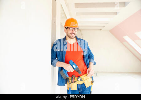 Portrait of young repairman wearing safety helmet and tool belt and using hammer drill driver while working on construction site. Stock Photo
