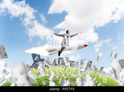 Conceptual image of young businessman in suit flying on rocket among flying papers with cityscape and blue sky on background. Stock Photo